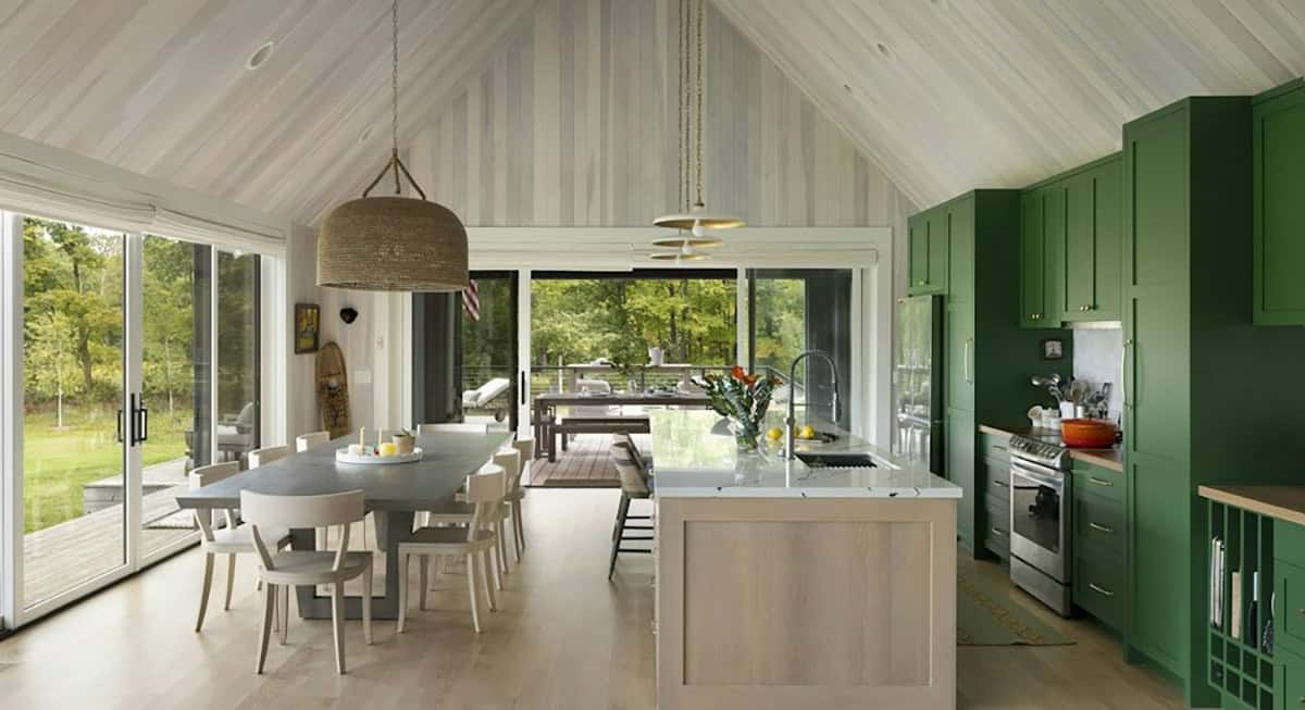 Dining table and white oak kitchen island beside green cabinets, looking through glass doors to a covered porch