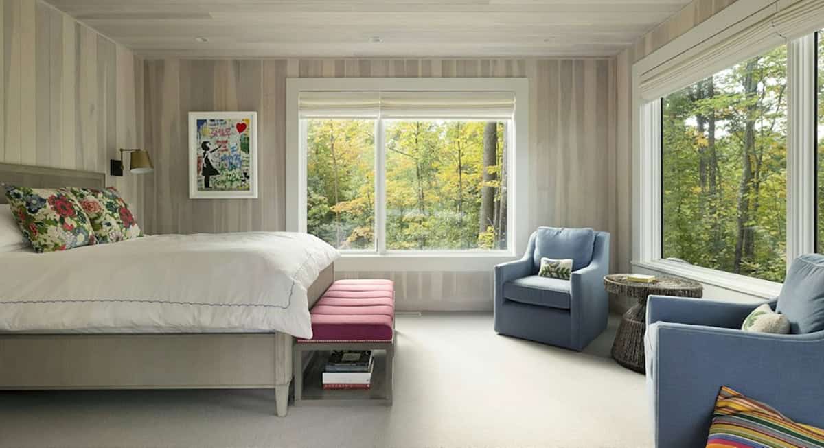 Primary bedroom with whitewashed wood walls, floral pillows, a pink bench and two blue swivel chairs by corner windows to the woods