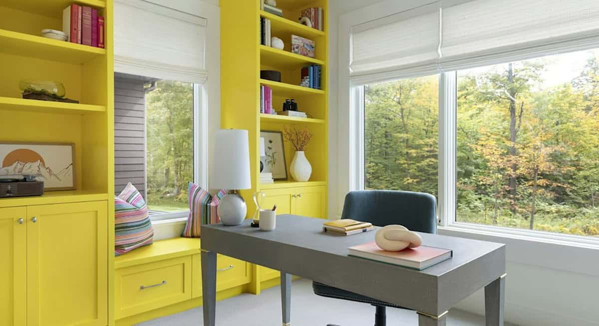 Home office with bright yellow built-in bookshelves and window seat, a gray desk and windows to fall trees