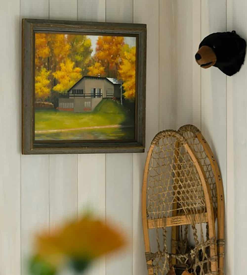Framed painting of a lakeside cabin in fall trees beside vintage snowshoes and a plush bear head on a whitewashed wall
