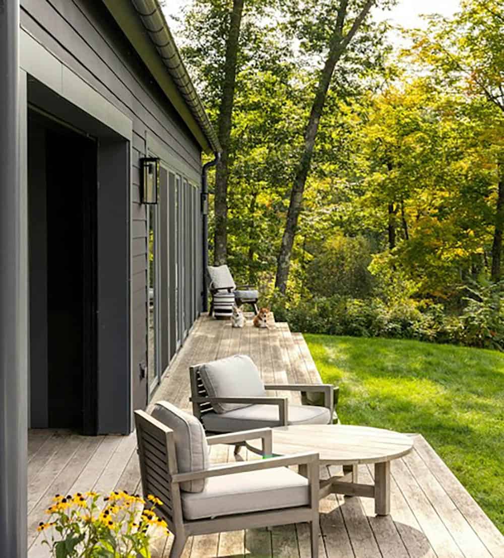 Wood deck with gray cushioned lounge chairs and a round table along the dark gray cabin, with two corgis resting at the far end