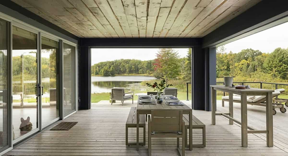Covered porch with a weathered wood ceiling, an outdoor dining table and benches, and a wide opening framing the lake