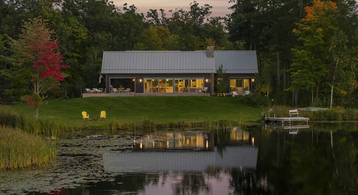 Dark gray lake cabin glowing at dusk across a lily pad lake, with yellow Adirondack chairs on the lawn and a small dock