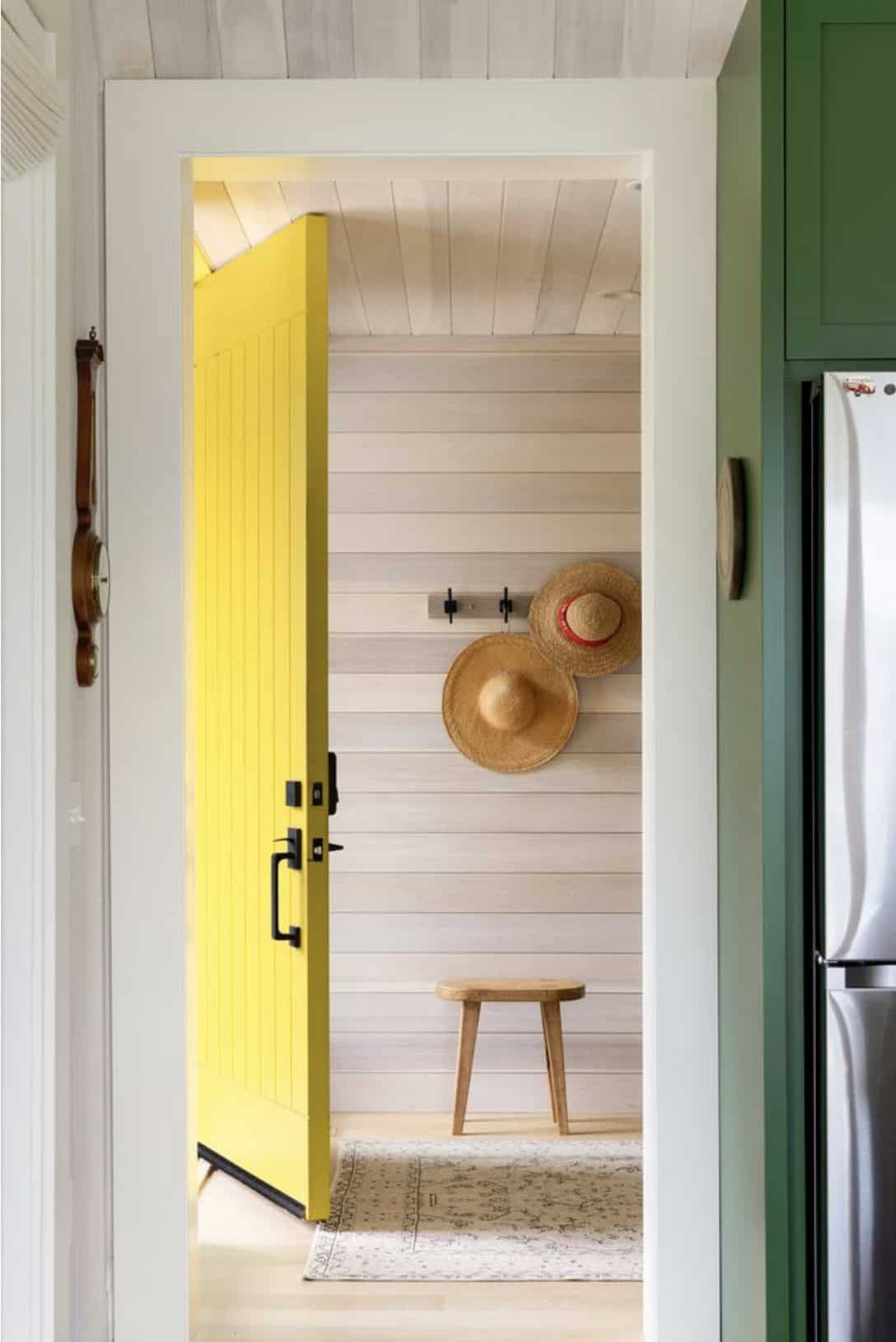 Bright yellow plank door open to a whitewashed entry with straw hats, a wooden stool and a vintage rug, beside green kitchen cabinets