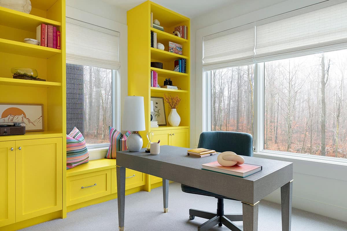Home office with bright yellow built-ins, a window seat with striped pillows and a gray desk facing a misty winter forest