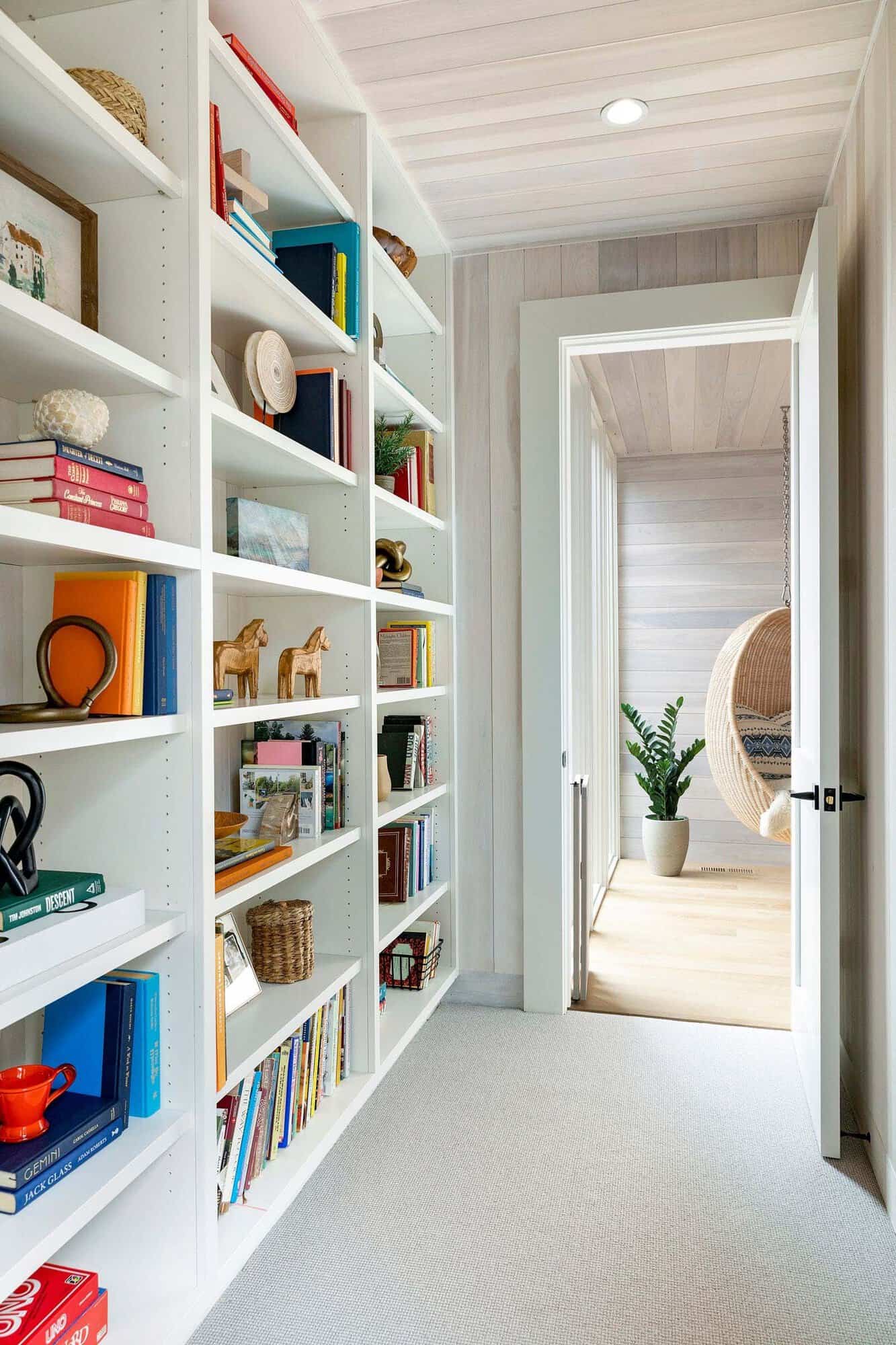 Hallway lined with white built-in bookshelves full of colorful books and decor, leading to a room with a hanging rattan chair