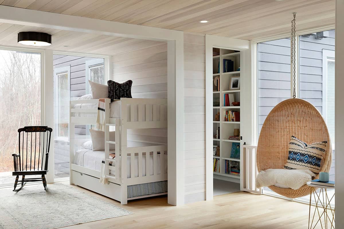 Bunk alcove with white bunk beds and a trundle, a black rocking chair and a hanging rattan egg chair by tall windows
