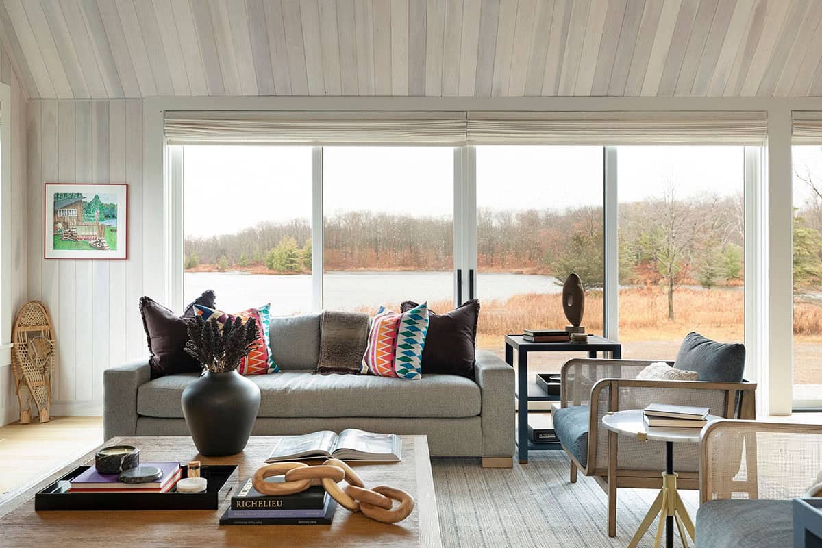 Living room with a gray sofa and colorful patterned pillows in front of a wall of glass overlooking the lake in late fall