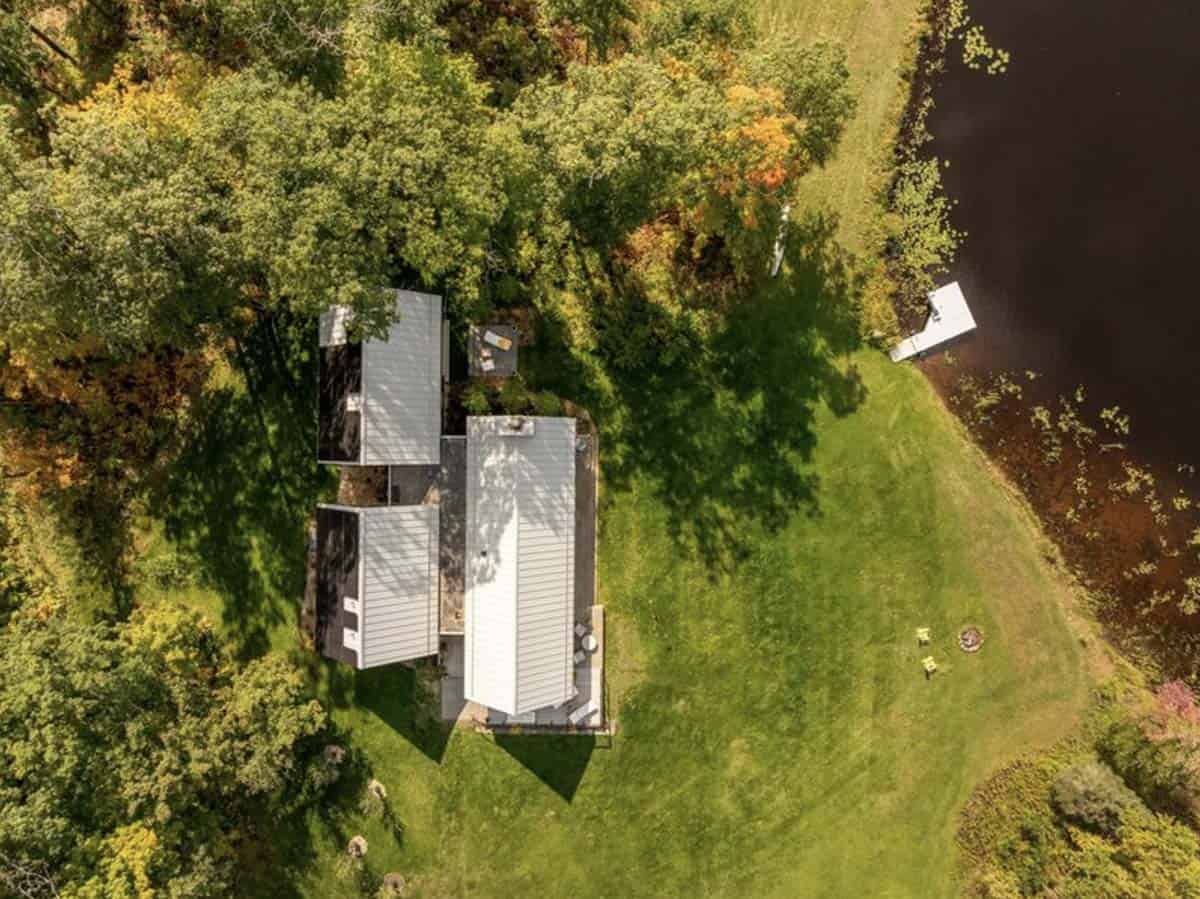 Aerial view of the cabin's three metal-roofed gables beside a green lawn that slopes down to the lake and a small dock