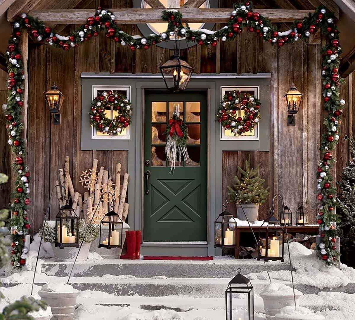 Rustic barn wood porch with a green door, red and white ornament garland, window wreaths and black lanterns