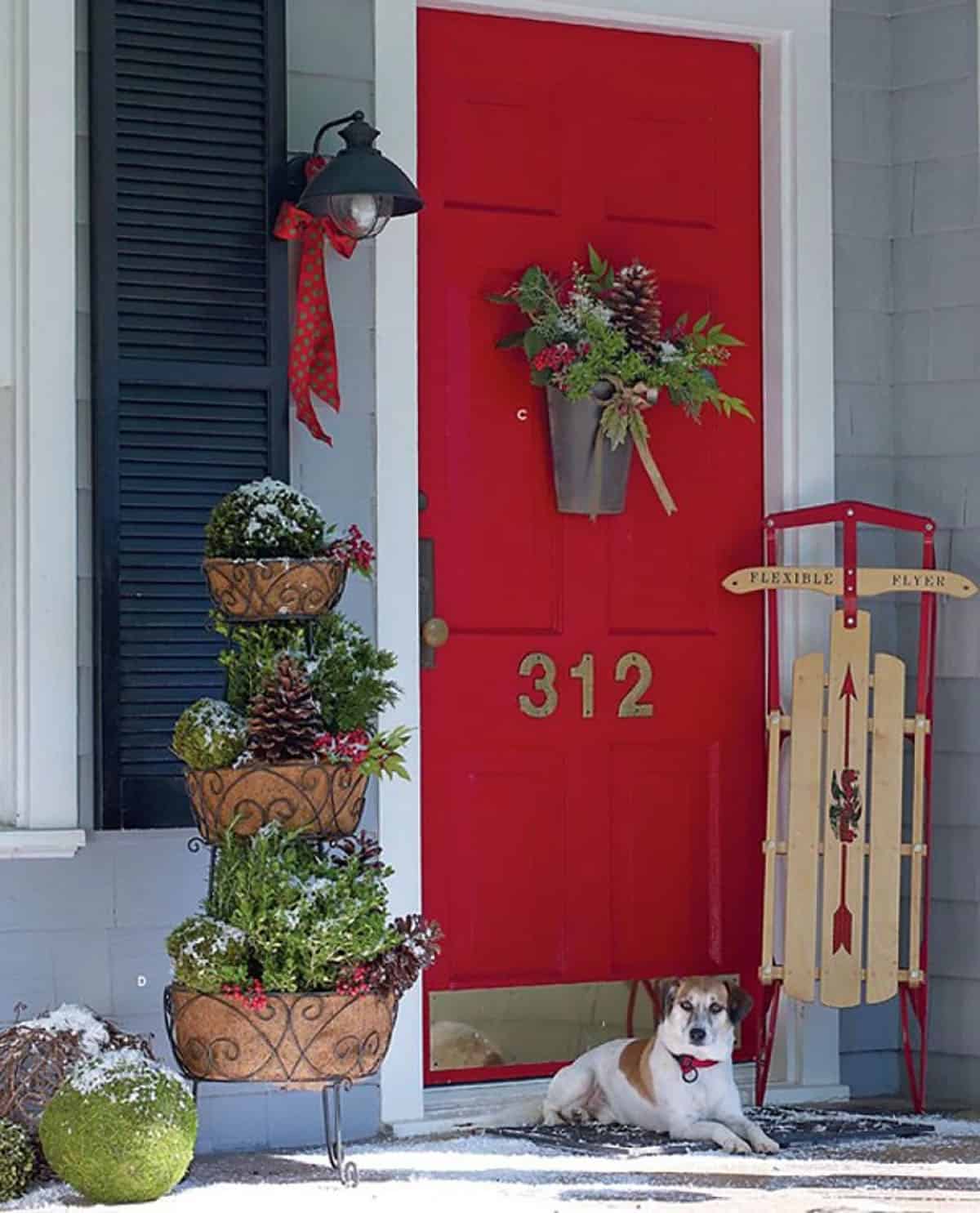 Red front door with a hanging bucket of greenery, a tiered evergreen planter and a vintage sled