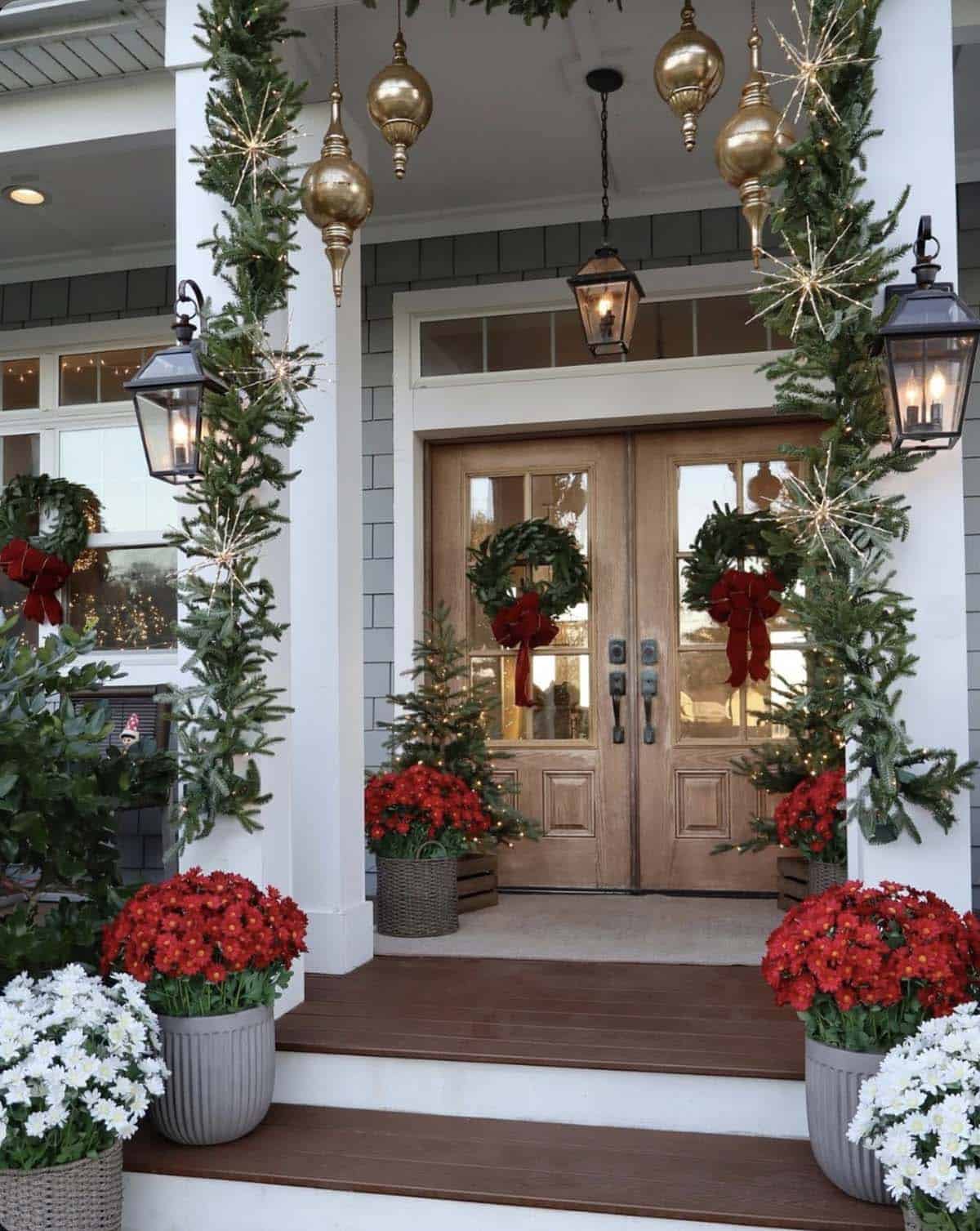 Front porch with garland-wrapped columns, hanging brass finial ornaments, wreaths with red bows on wood double doors and pots of red and white mums on the steps