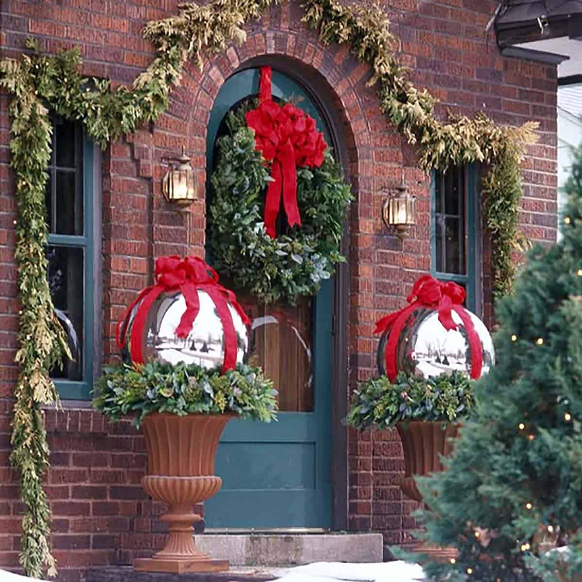 Teal arched front door with a red-bowed wreath and giant silver ornaments in urns on a brick house