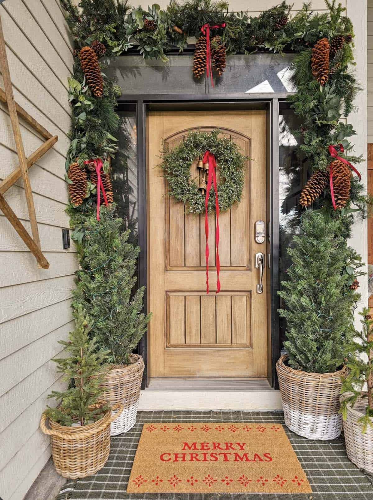 Wood front door framed by evergreen garland with large pinecones and red ribbons, a wreath, potted trees in woven baskets and a Merry Christmas doormat