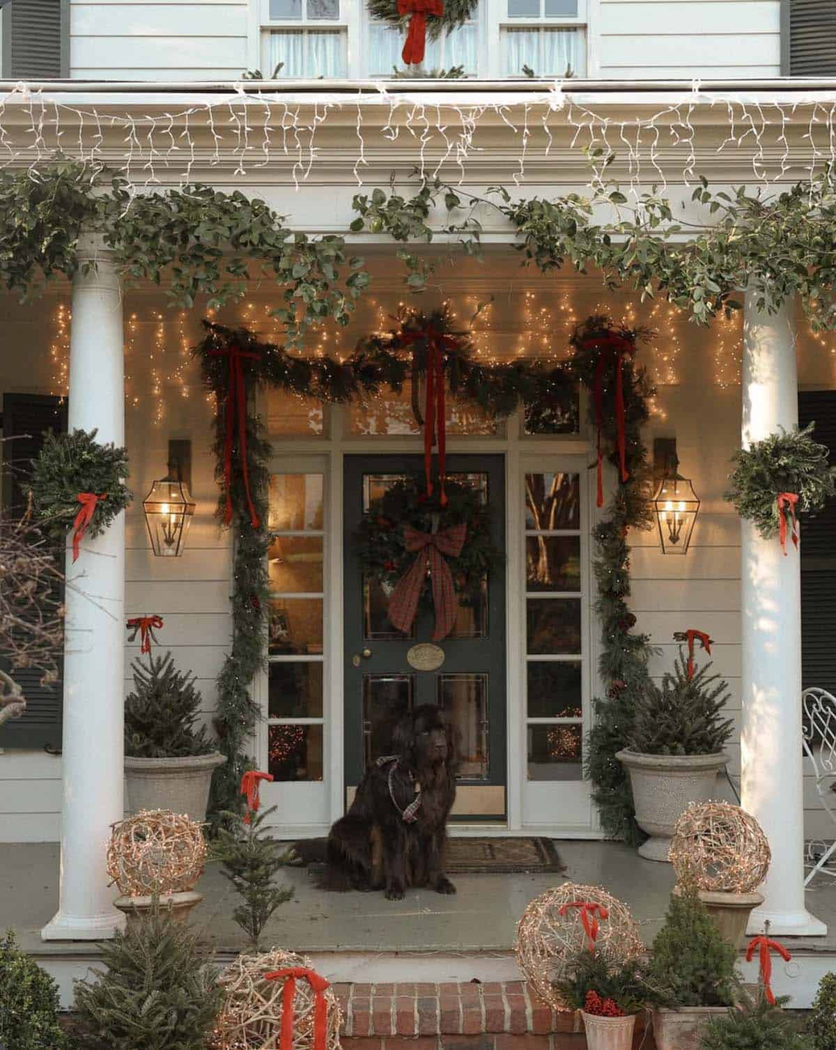 White porch with icicle lights, cedar garland tied with red velvet ribbons, a plaid bow wreath on a dark door, lighted grapevine orbs and a dog on the porch