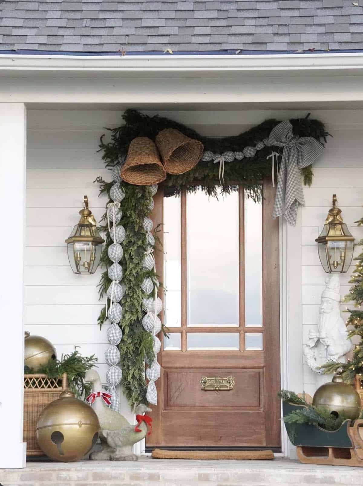Wood front door with cedar garland, woven bells, a blue gingham fabric ball garland and bow, giant brass jingle bells and stone geese