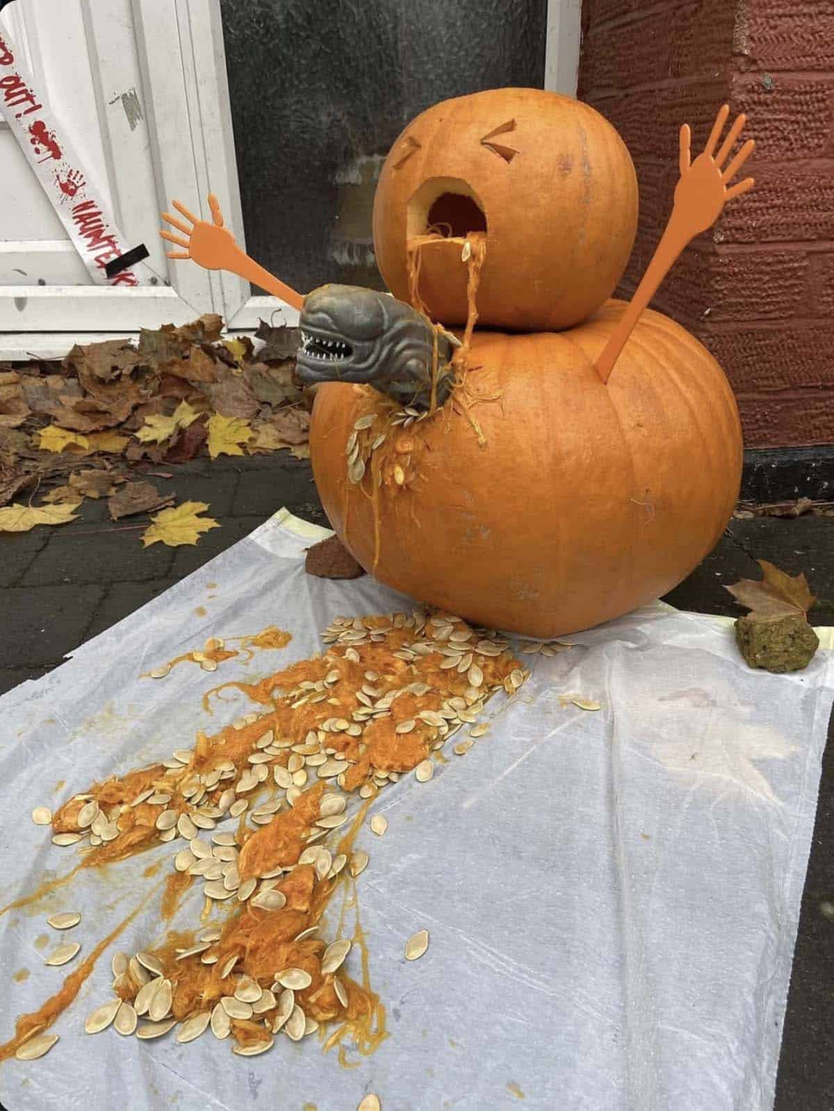 two stacked pumpkins with a screaming carved face and plastic hands, with a toy alien bursting out of the bottom pumpkin and seeds spilling on the ground