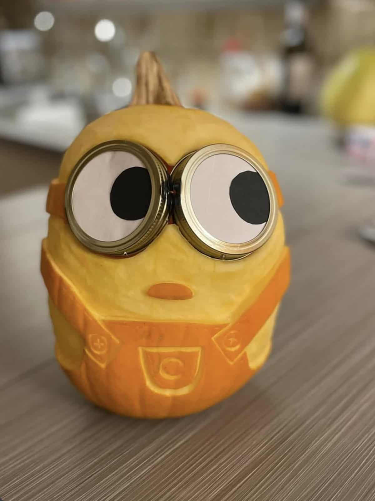 pumpkin carved like a Minion with mason jar lid goggles, googly eyes, a small O-shaped mouth and carved overalls