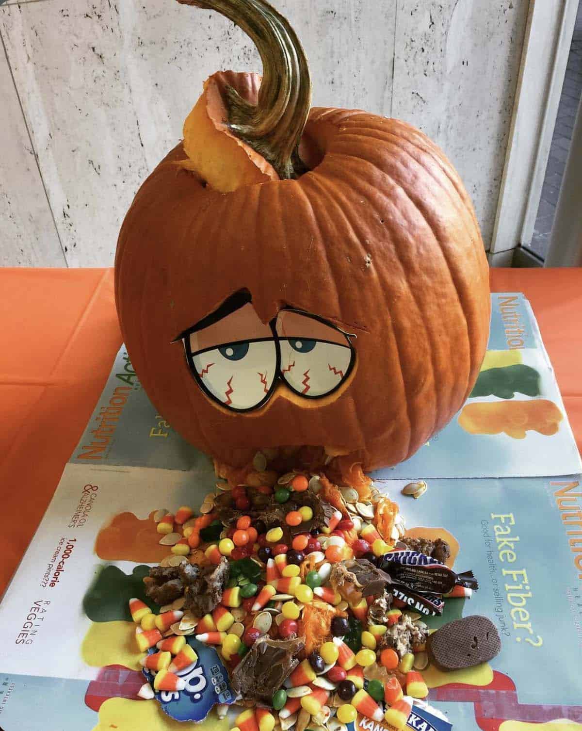 carved pumpkin with droopy bloodshot cartoon eyes, spilling candy corn, chocolate bars, candies and pumpkin seeds from its mouth