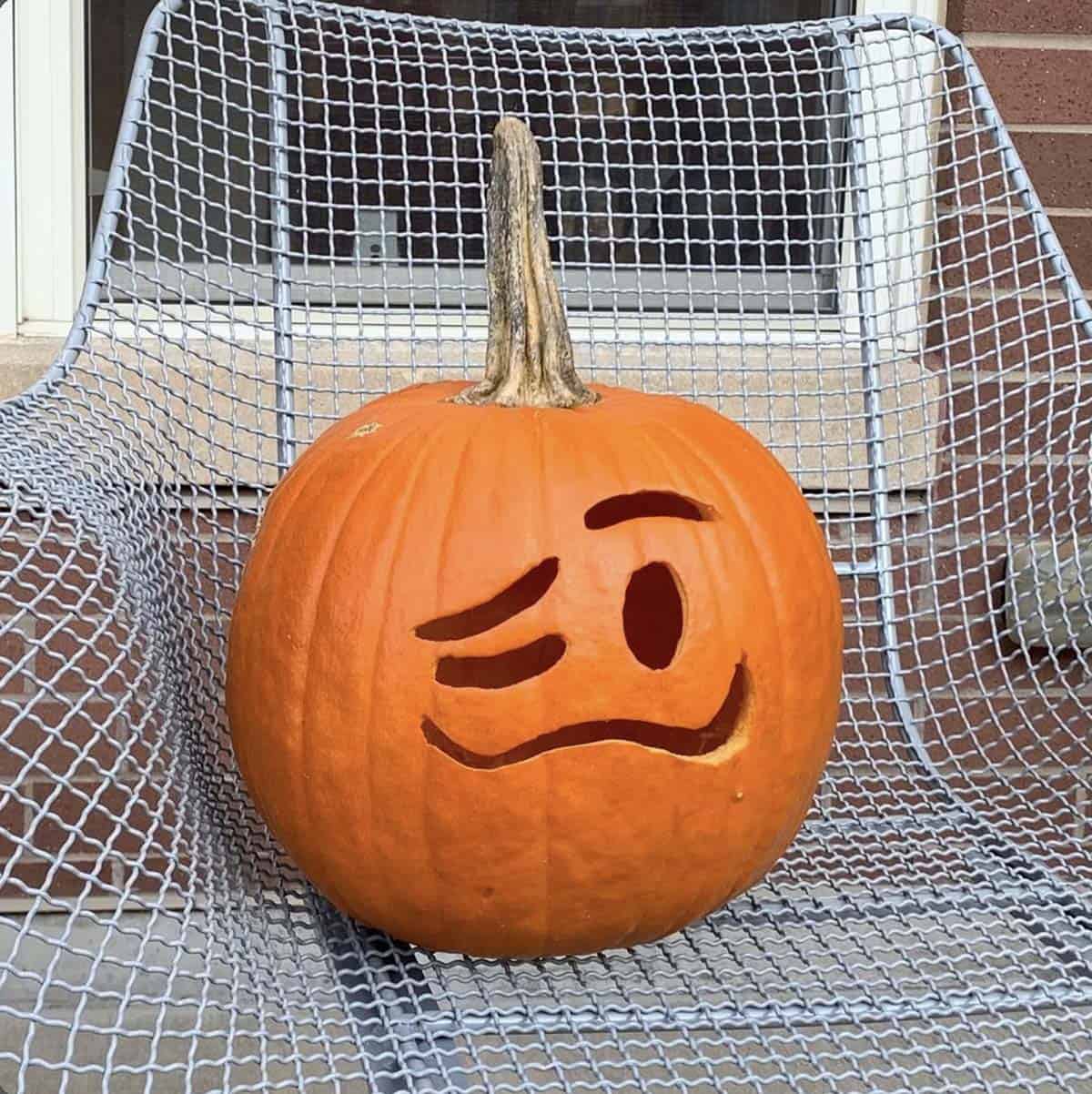 pumpkin carved like the woozy face emoji, with one squinting eye, one wide eye and a wavy lopsided grin