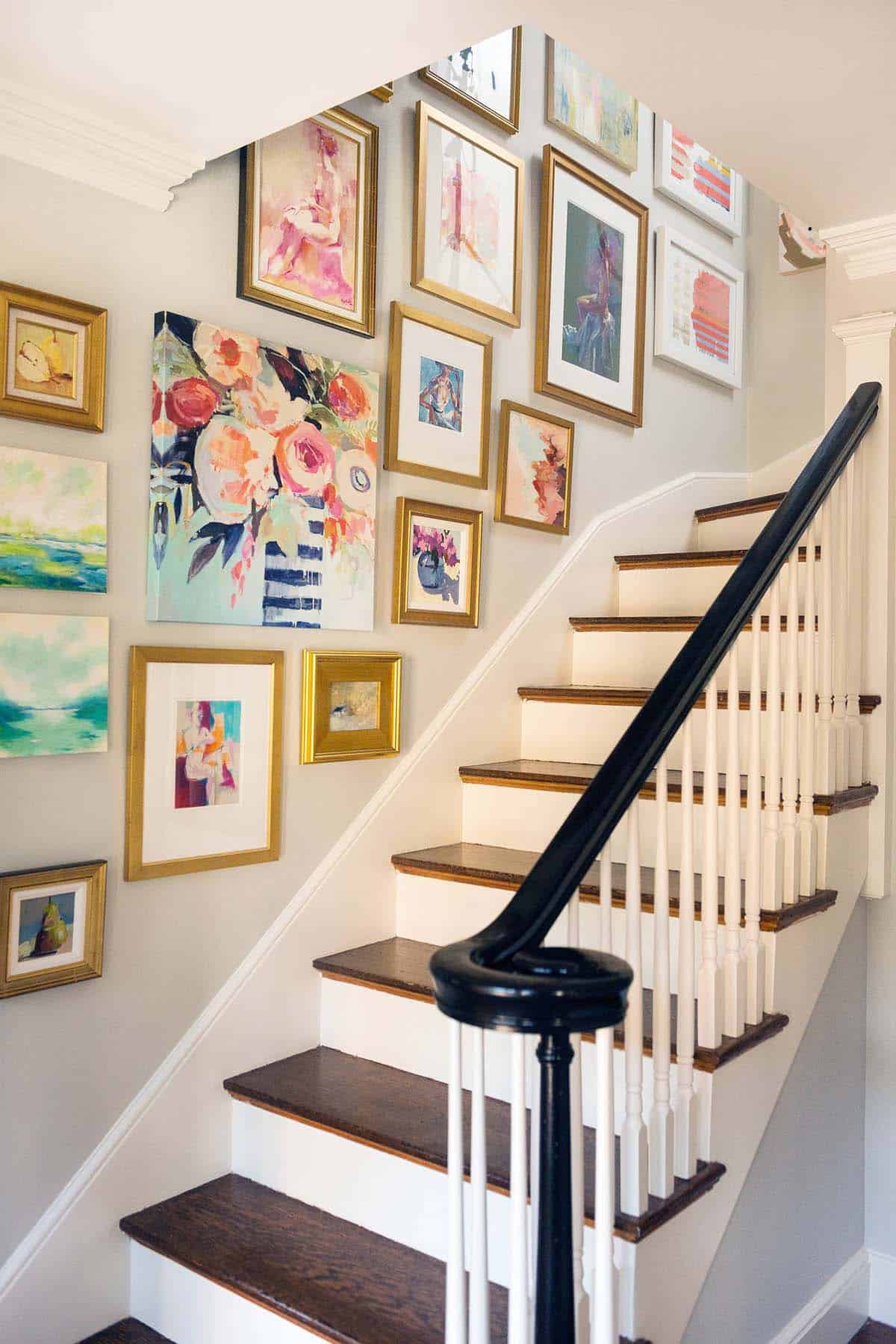 Staircase gallery wall of colorful paintings in gold frames alongside white stairs with a black handrail