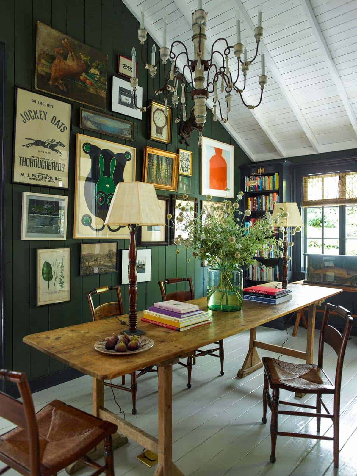 Eclectic gallery wall of vintage posters and paintings on a dark green paneled wall above a rustic dining table