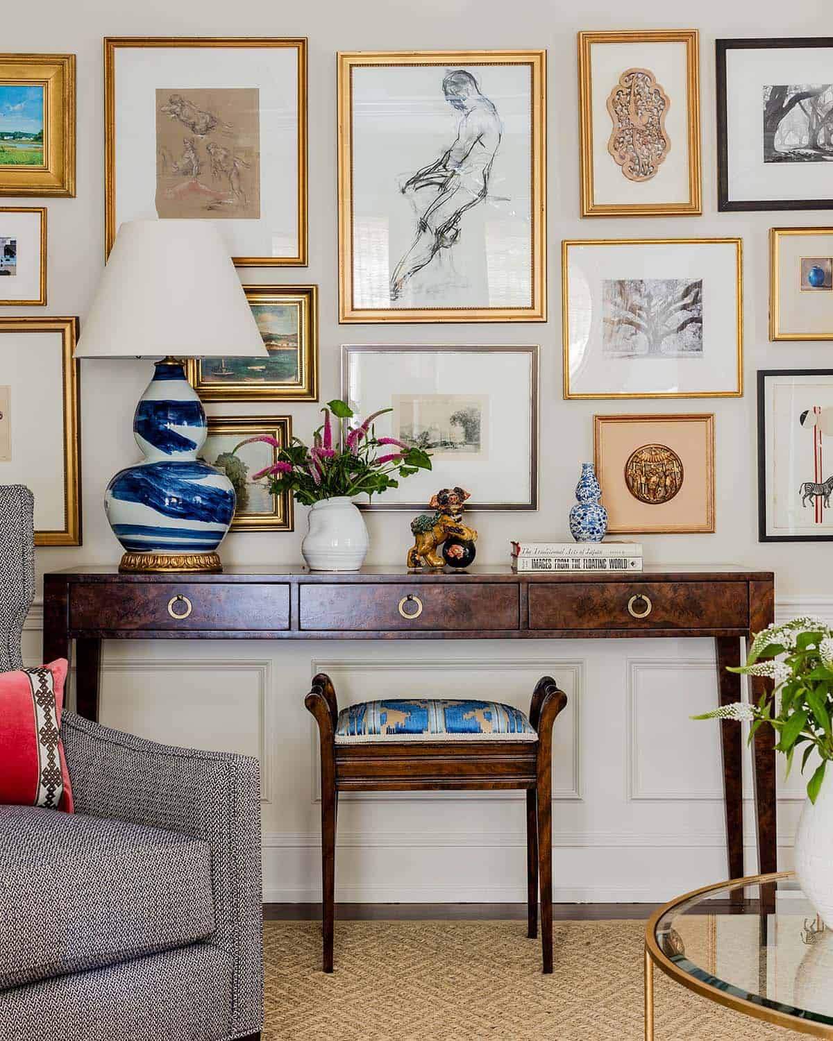 Collected gallery wall of sketches and paintings in gold frames above a burl wood console table