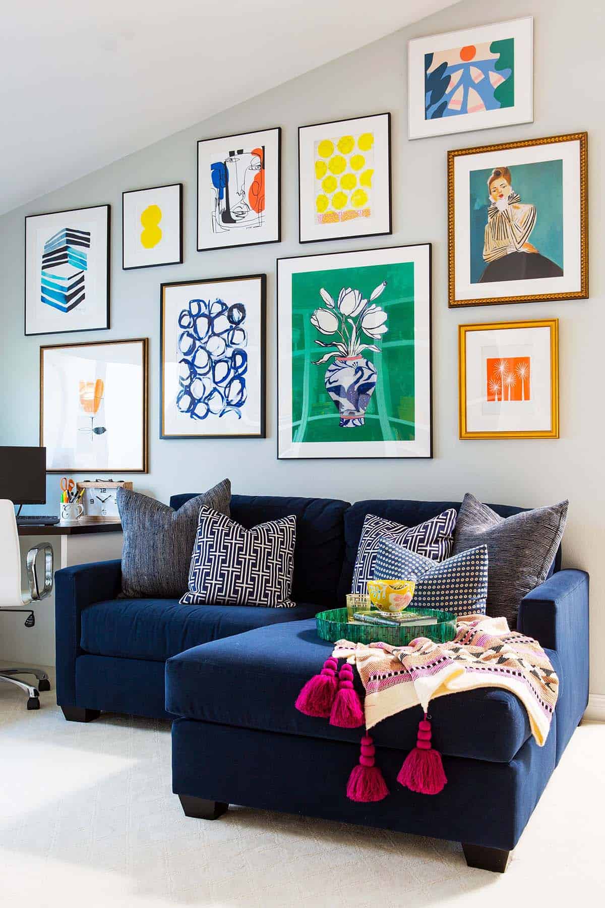 Colorful gallery wall of framed art prints under a sloped ceiling above a navy sectional in a home office