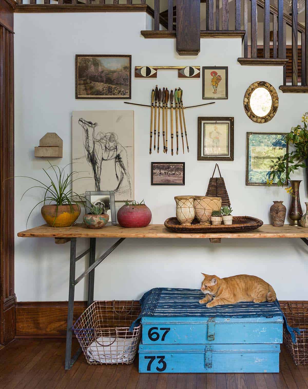 Eclectic gallery wall of vintage art, antique arrows and a shell mirror above a rustic console beneath a staircase