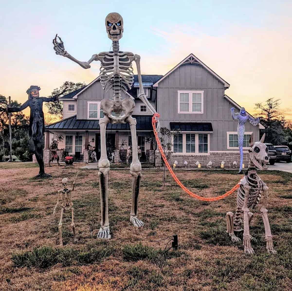 Giant skeleton holding an orange chain leash to a skeleton dog on a lawn with a scarecrow, ghost, and mini skeleton at sunset