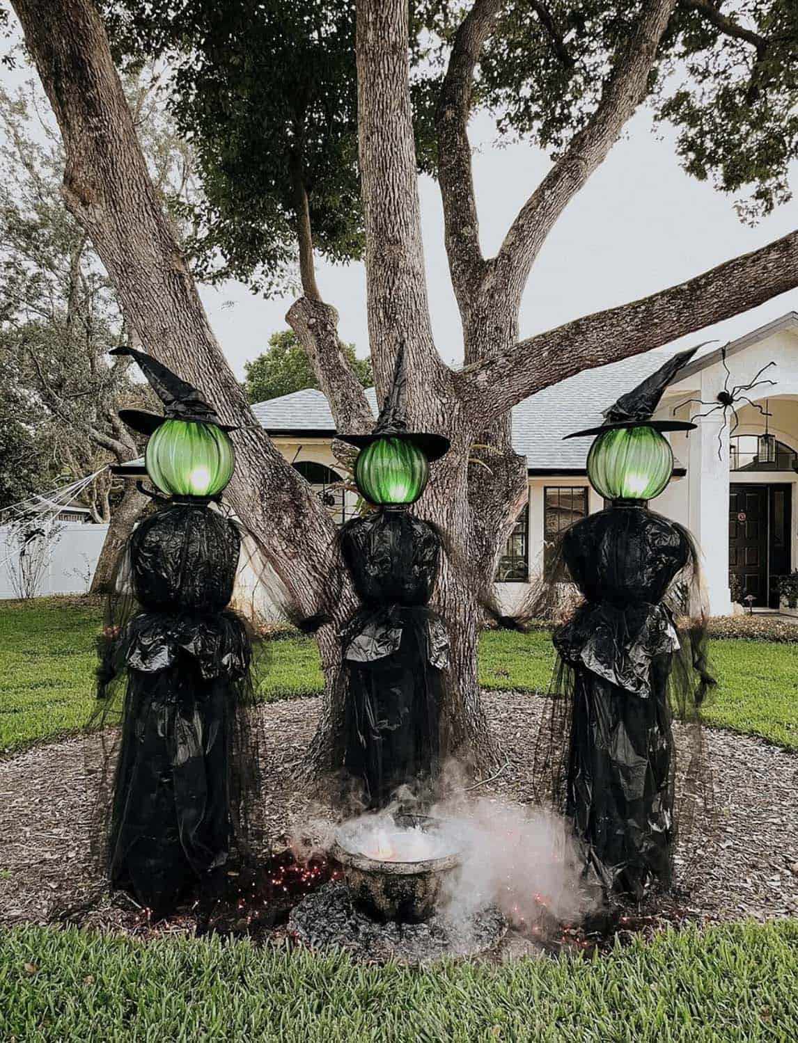 Three DIY witches with glowing green heads, black trash bag dresses, and pointed hats around a smoking cauldron under a tree