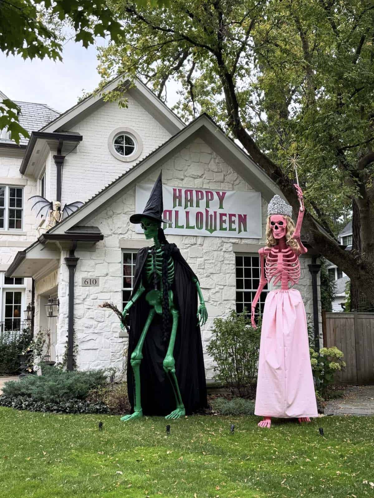 Giant green witch skeleton and pink good witch skeleton in a gown and crown on a lawn under a Happy Halloween banner