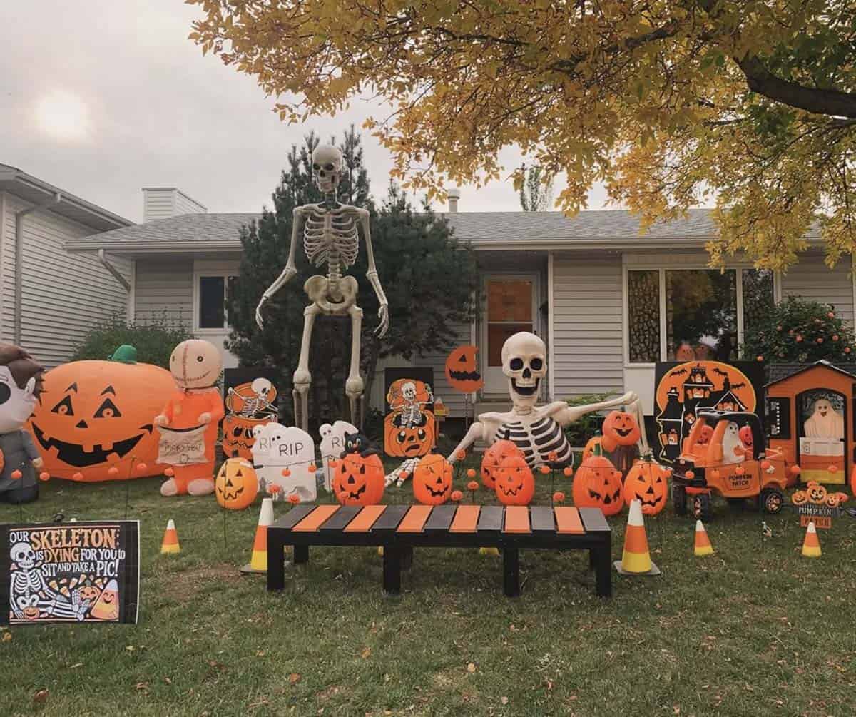 Halloween photo op lawn with giant skeleton, striped bench, glowing pumpkins, tombstones, candy corn cones, and a sit-and-take-a-pic sign