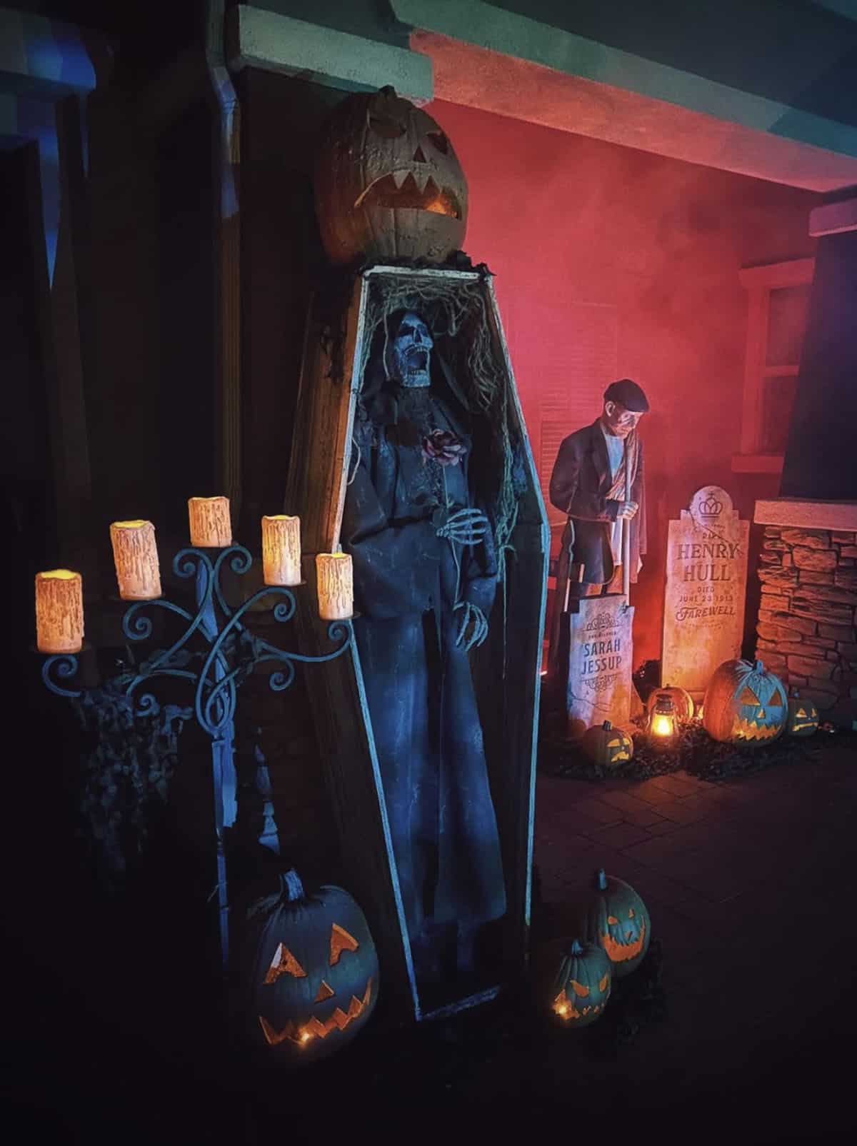 Upright coffin with a hooded corpse holding a rose, topped with a jack-o'-lantern, beside a candelabra, tombstones, and a gravedigger in red fog