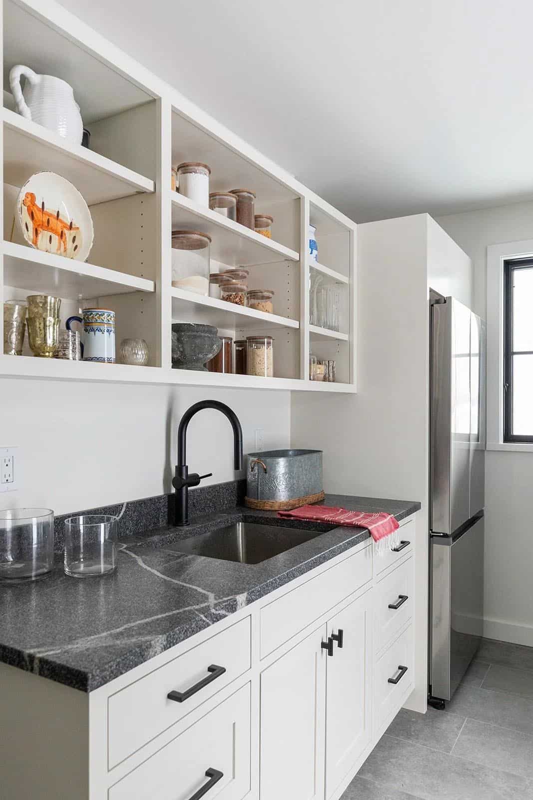 Pantry with open white shelving, dark soapstone counter, black faucet and stainless refrigerator