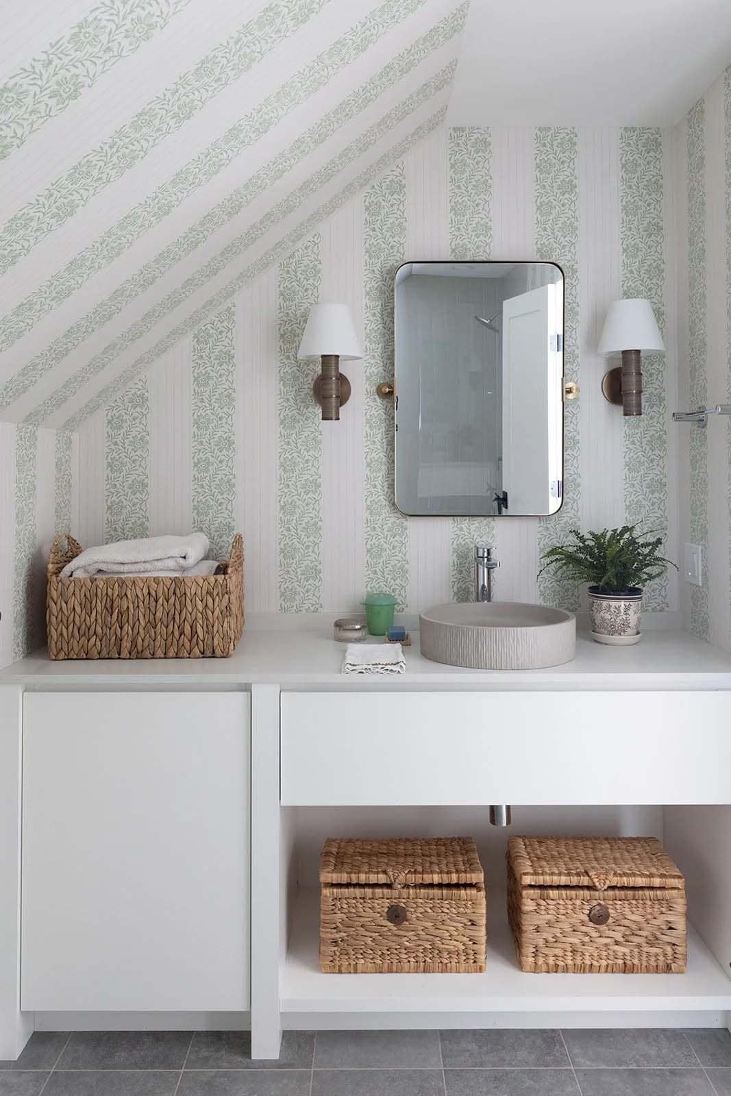 Vanity with woven baskets, two sconces and striped wallpaper under a sloped ceiling