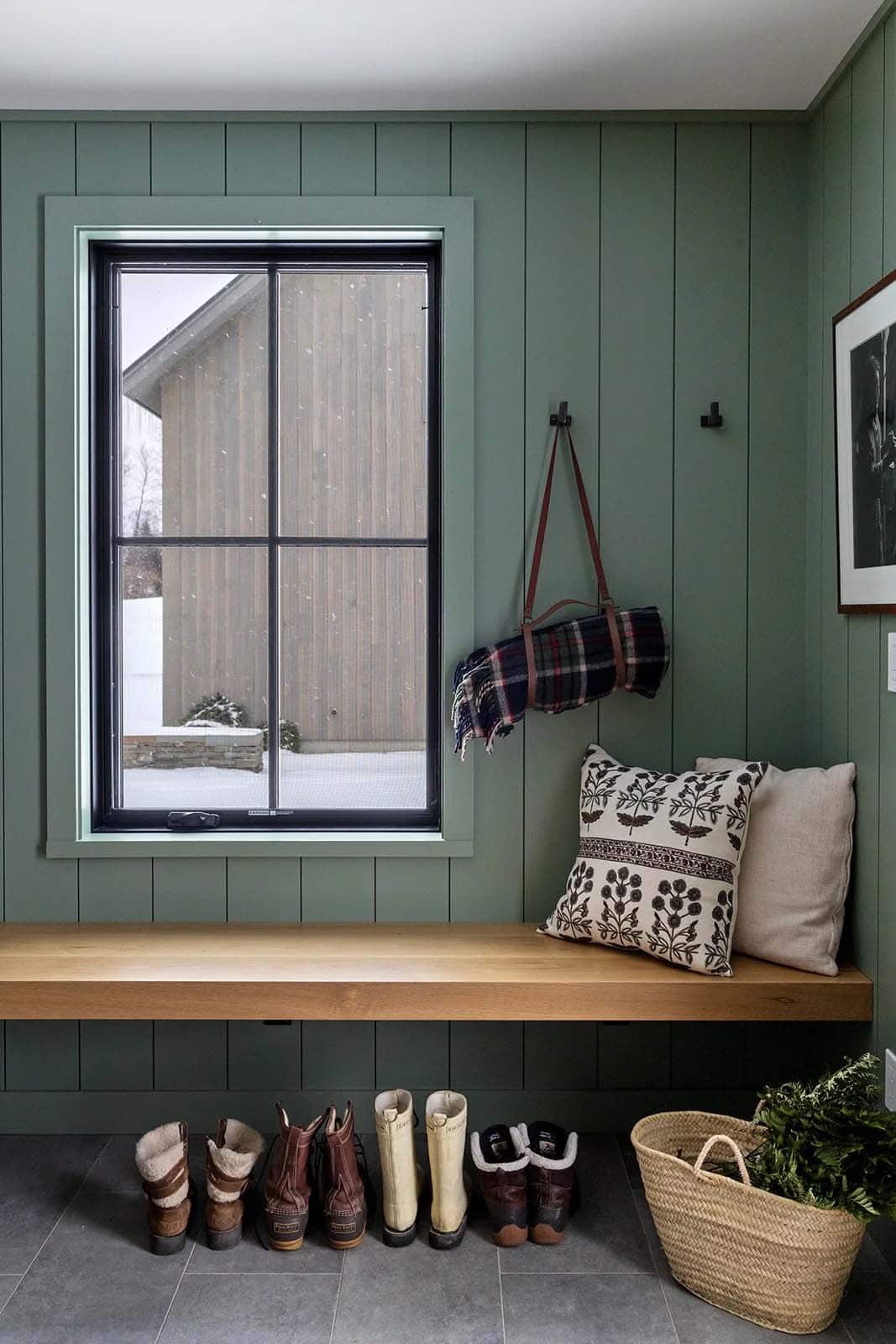 Mudroom with sage green paneling, a floating oak bench, a plaid blanket and winter boots beneath a window