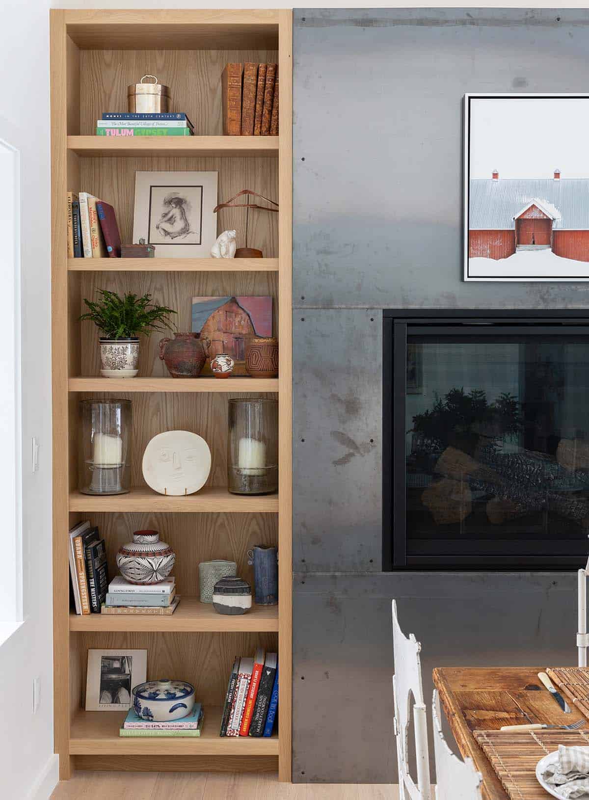 Oak bookshelf with curated pottery and books beside a steel fireplace and red barn artwork