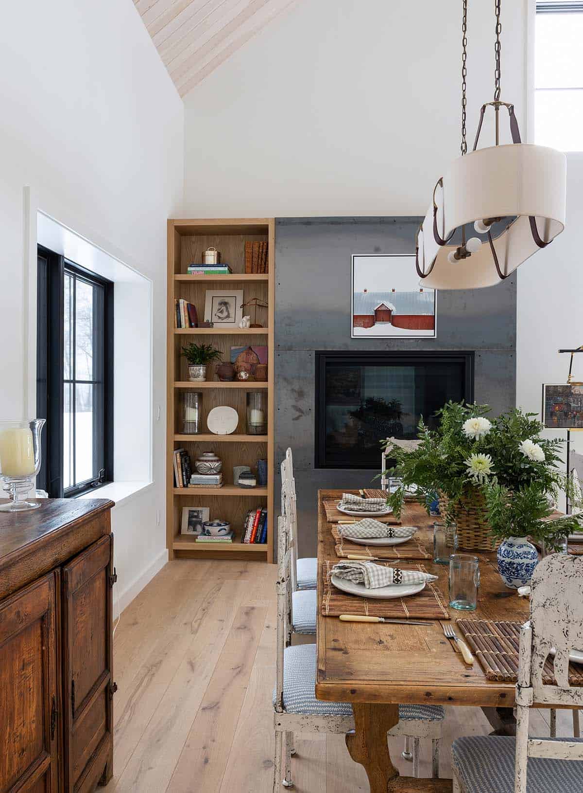 Dining area with a rustic table, bookshelf and steel fireplace beneath a vaulted ceiling