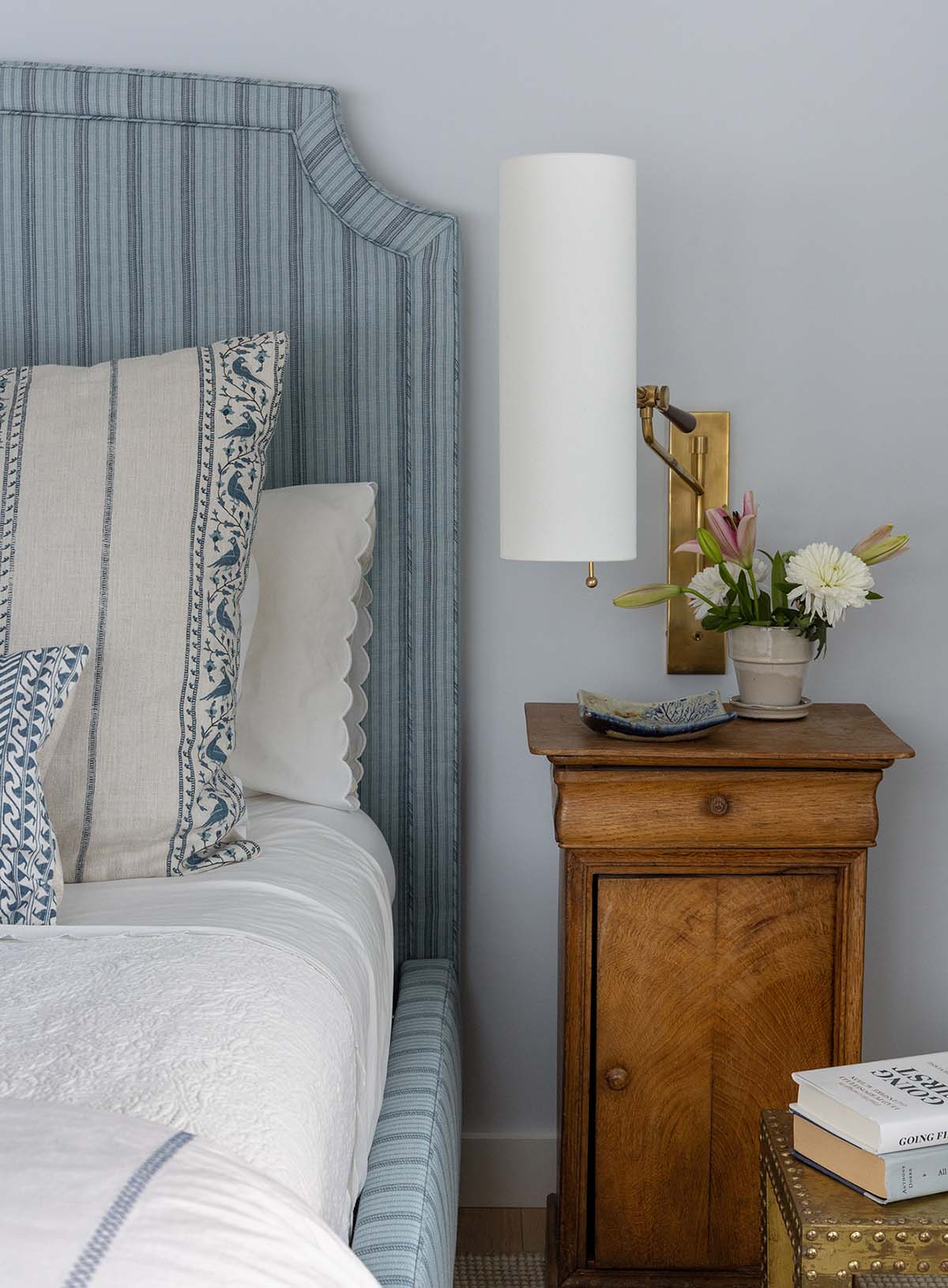 Bedroom with a blue striped upholstered headboard, antique wood nightstand and brass wall sconce