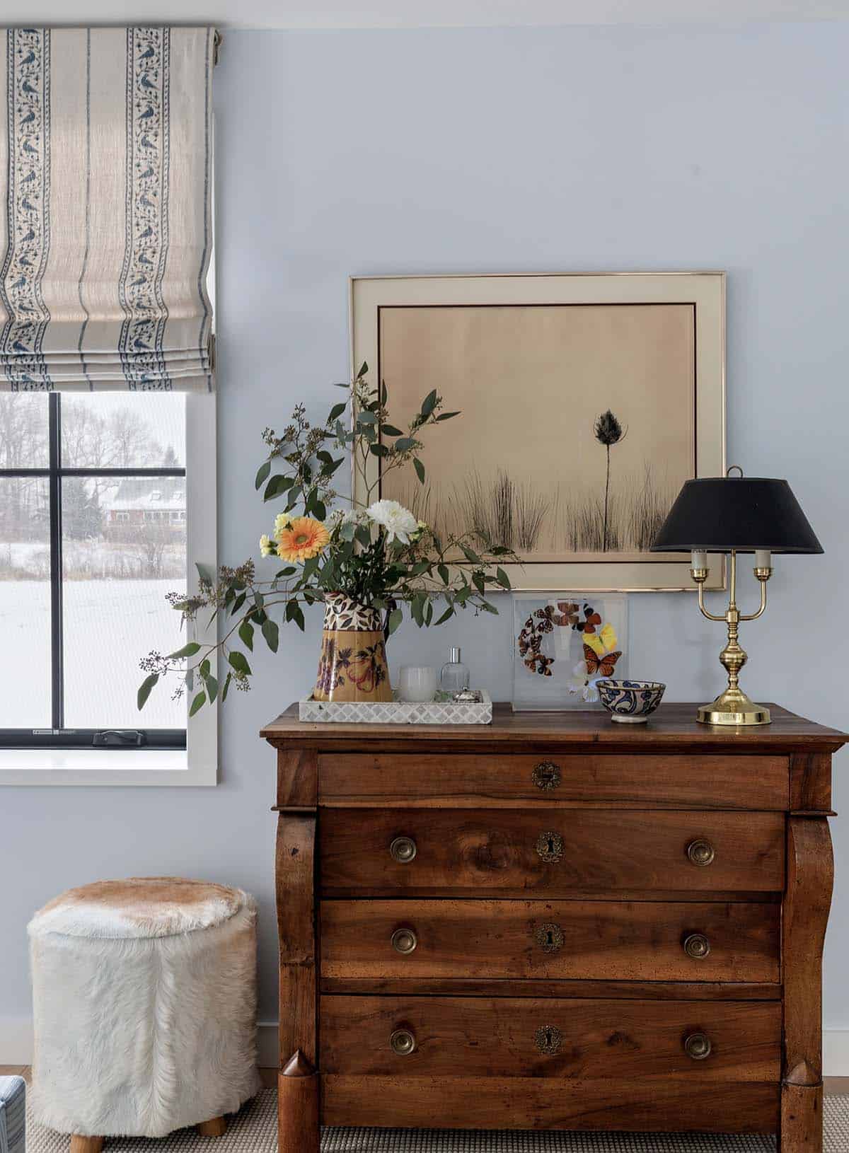 Antique wood dresser with a framed botanical print, brass lamp and cowhide stool beside a snowy window