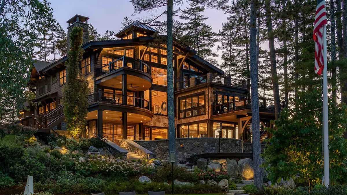 Timber frame lakefront lodge at dusk with curved balconies, stone chimney, and glowing windows