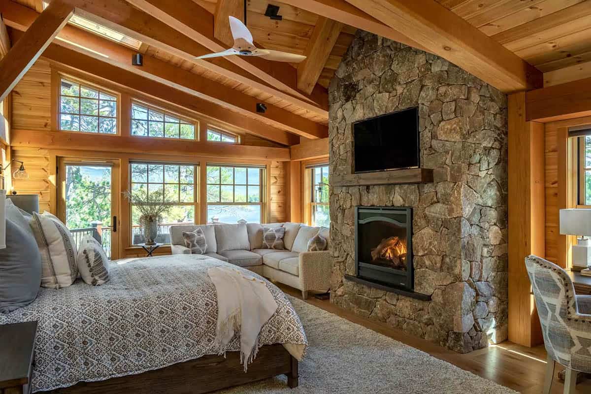 Primary bedroom with stone fireplace, vaulted beamed ceiling, and sectional facing lake views