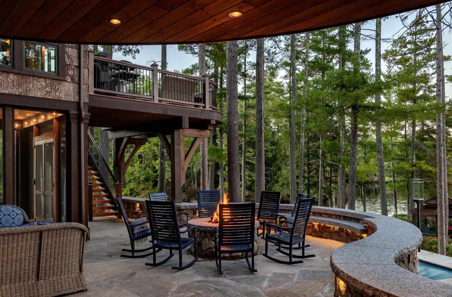 Stone terrace with fire pit and rocking chairs beneath a curved deck overlooking the lake