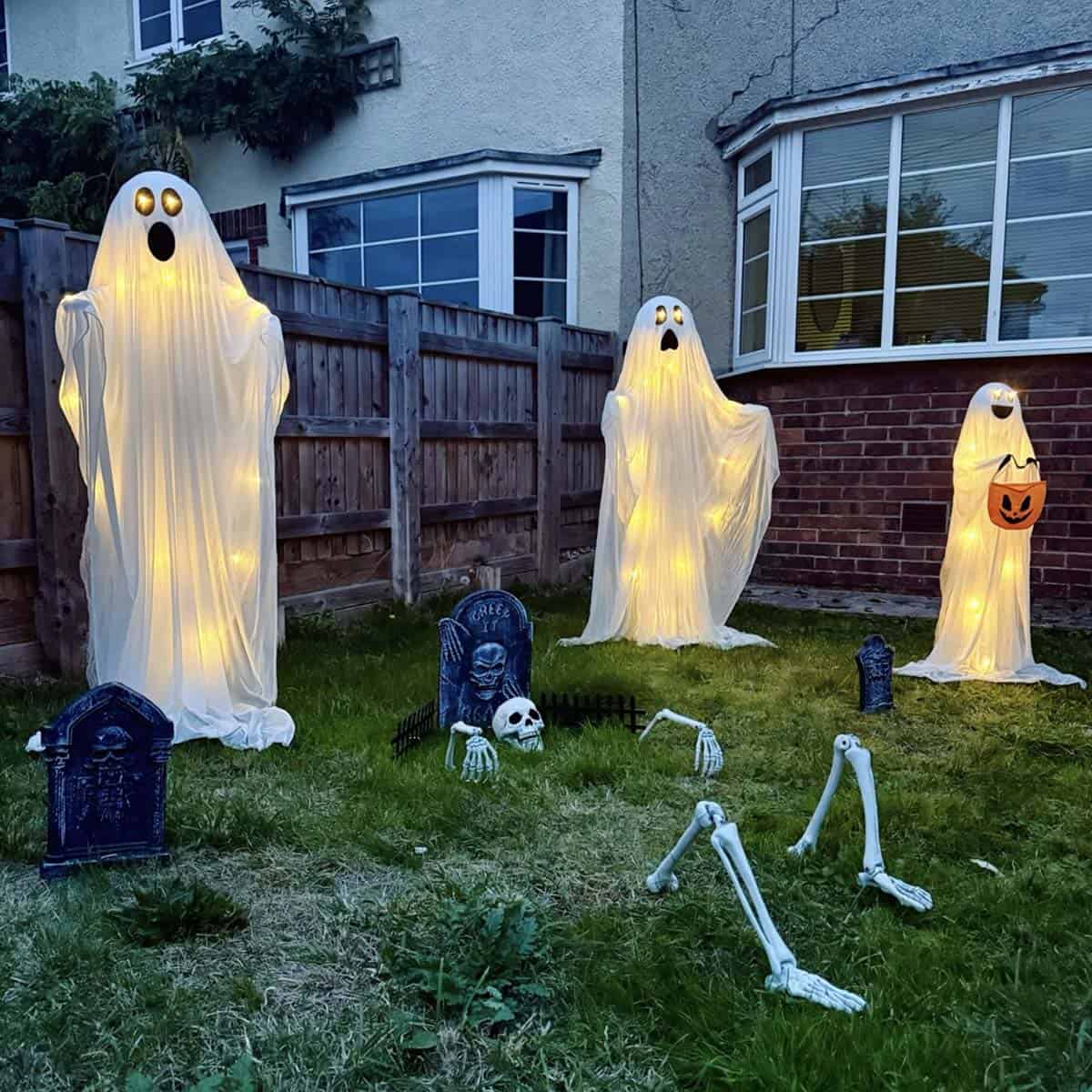 Three glowing ghosts with string lights, one holding a pumpkin bucket, beside tombstones and a skeleton rising from the lawn