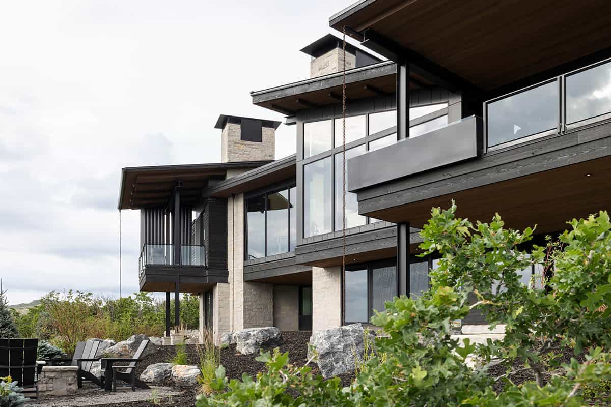 Rear exterior with dark wood siding, stone chimneys, and cantilevered decks above boulders
