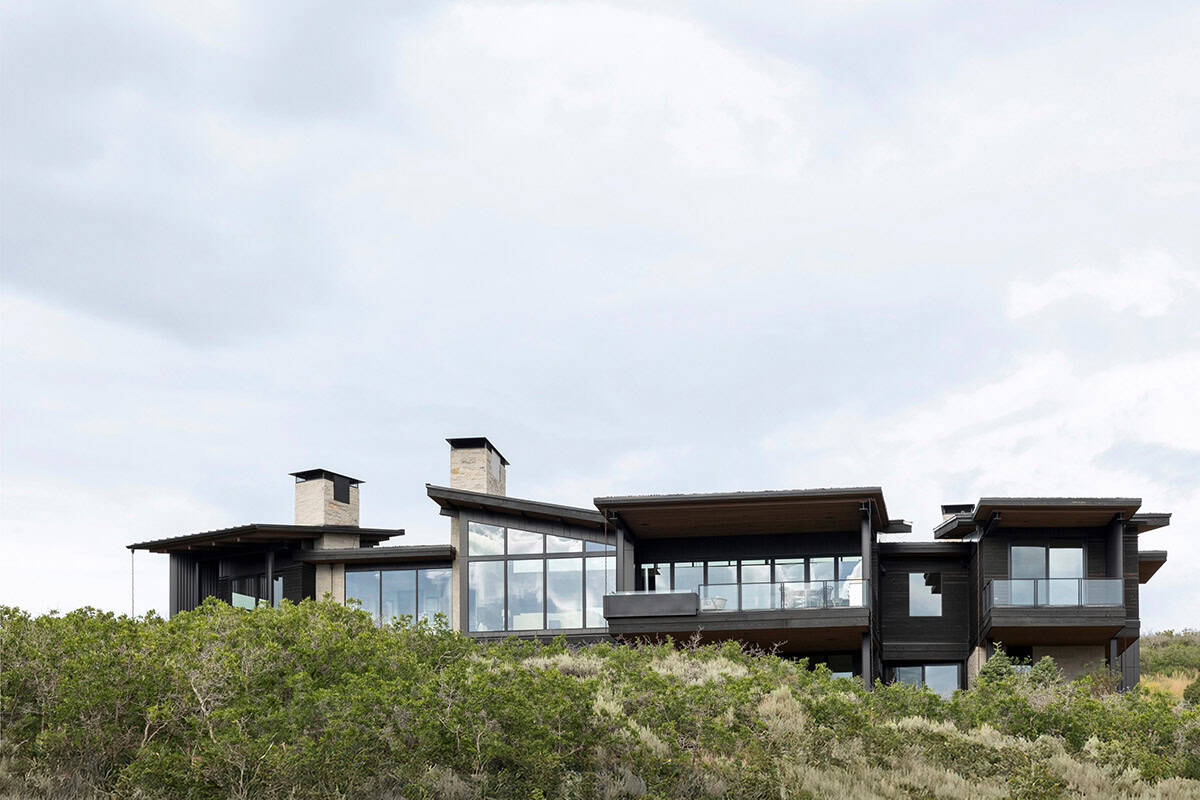 Dark-clad modern mountain home with stone chimneys rising above scrub oak