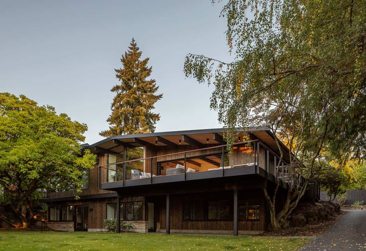 Mid-century modern home with cantilevered deck, dark wood siding, and mature trees at dusk