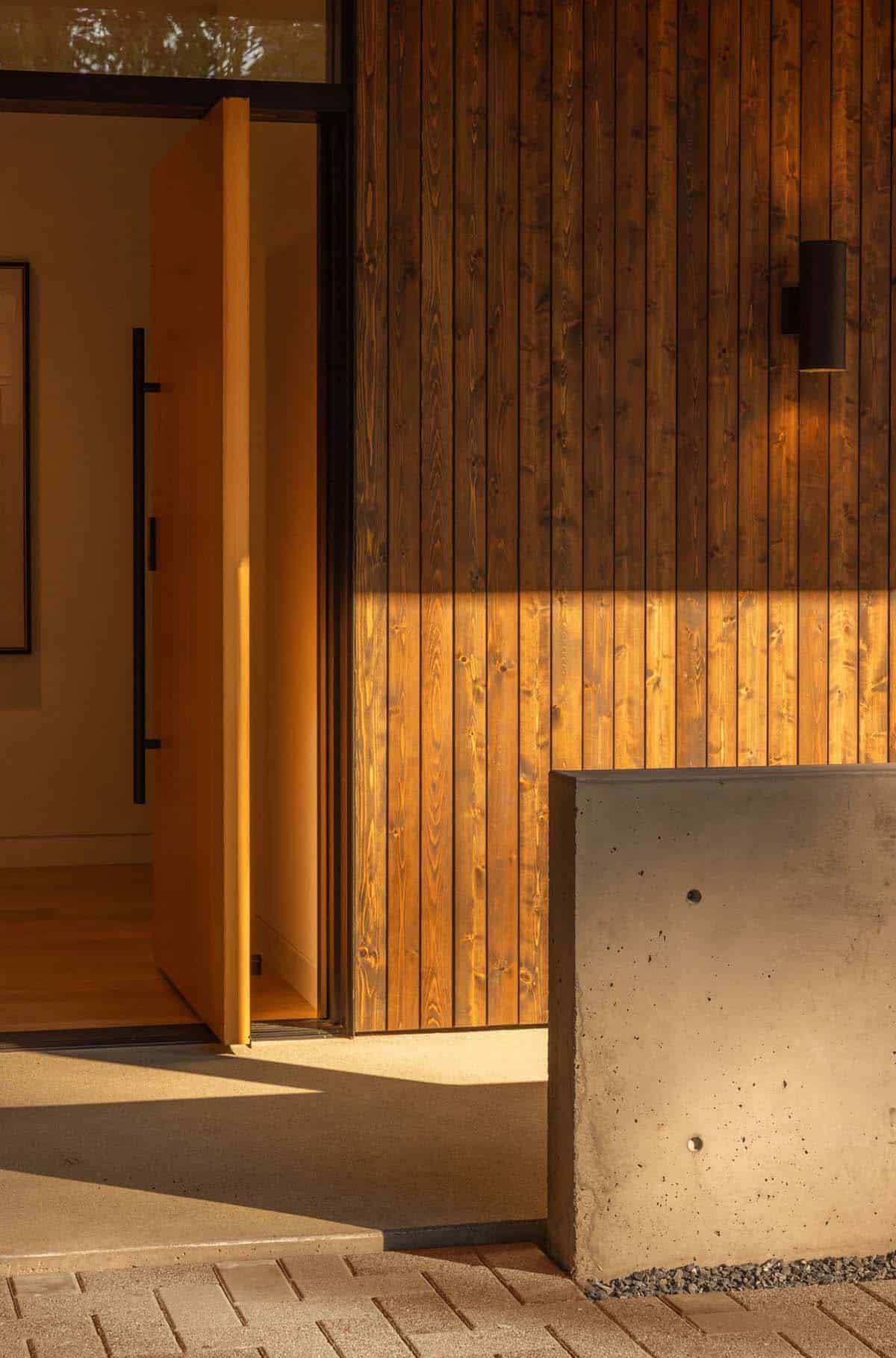 Open oak pivot front door beside stained cedar siding and concrete wall