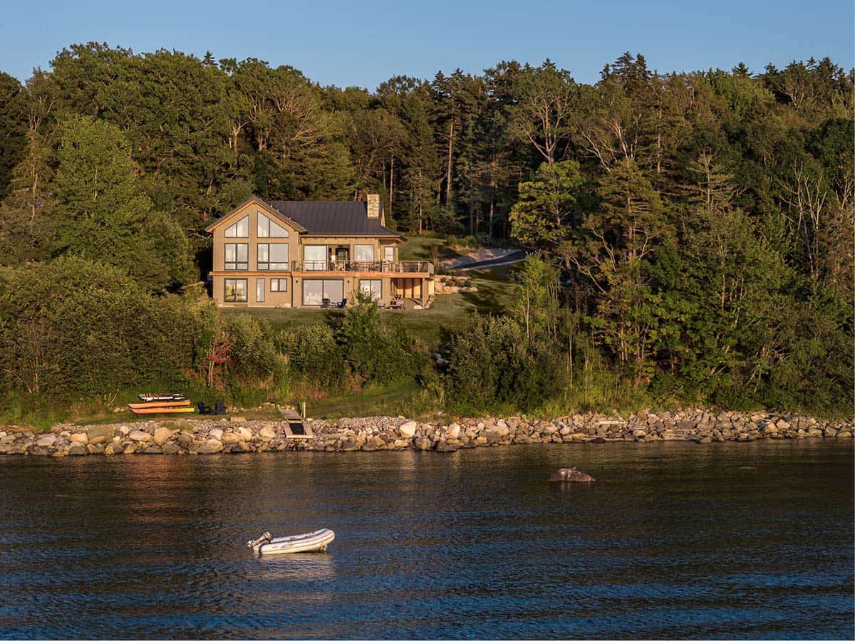Tan house with large windows and a dark metal roof above a rocky shoreline, with kayaks on the lawn and a small inflatable boat offshore